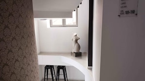 Un intérieur moderne avec un mur blanc, une fenêtre et une sculpture. On y trouve deux tabourets noirs et un panneau indiquant les numéros des chambres, du 101 au 110.