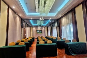 Un salón de conferencias con filas de mesas y sillas, una araña y una pantalla grande al frente.