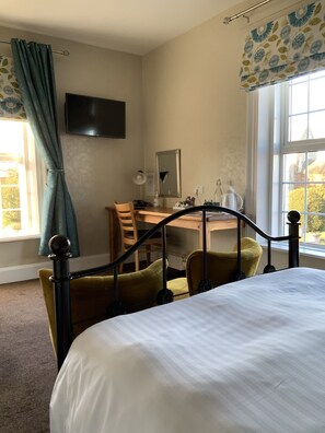 A bedroom with a bed, a desk, and two chairs. There is a window with curtains and a TV mounted on the wall.