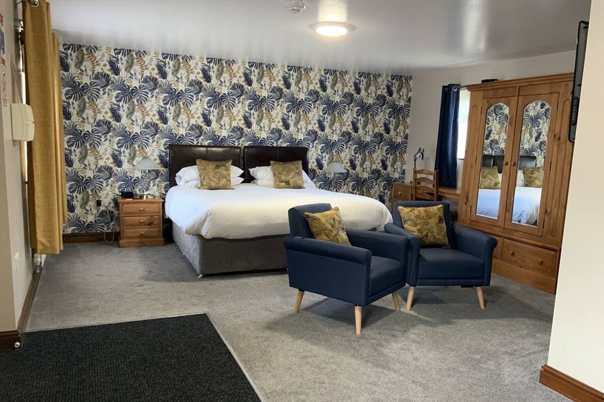 A bedroom with a bed, two armchairs, a wooden wardrobe, and a carpeted floor.