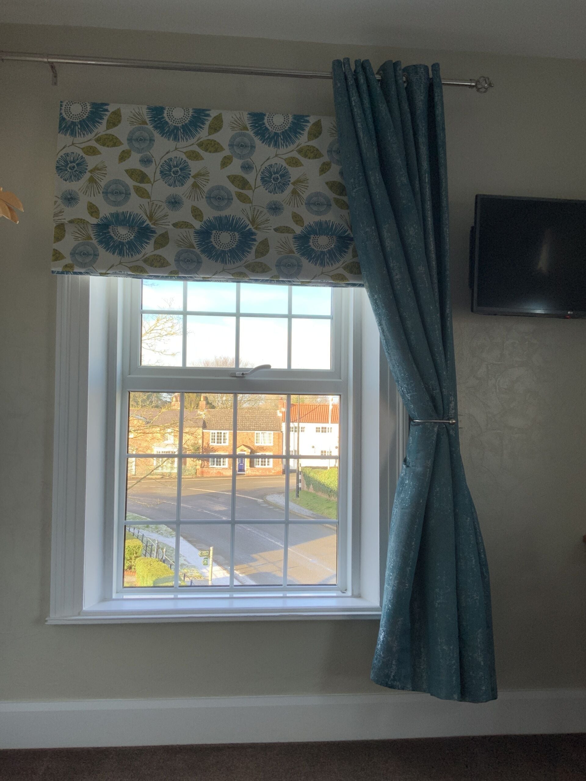 A window with a patterned valance and a curtain, a television on the wall, and a view of a street and buildings outside.