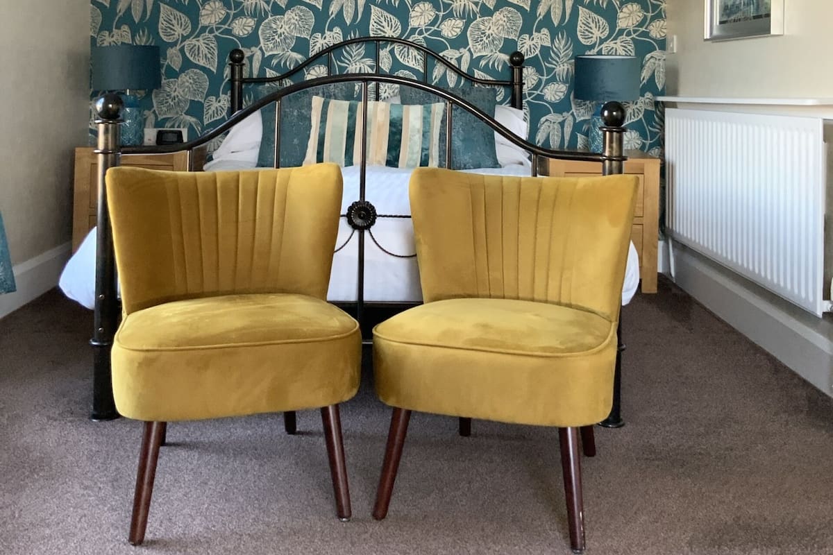 A bedroom with a floral wallpaper, a bed with a metal frame, two yellow chairs, a chandelier, and a radiator.