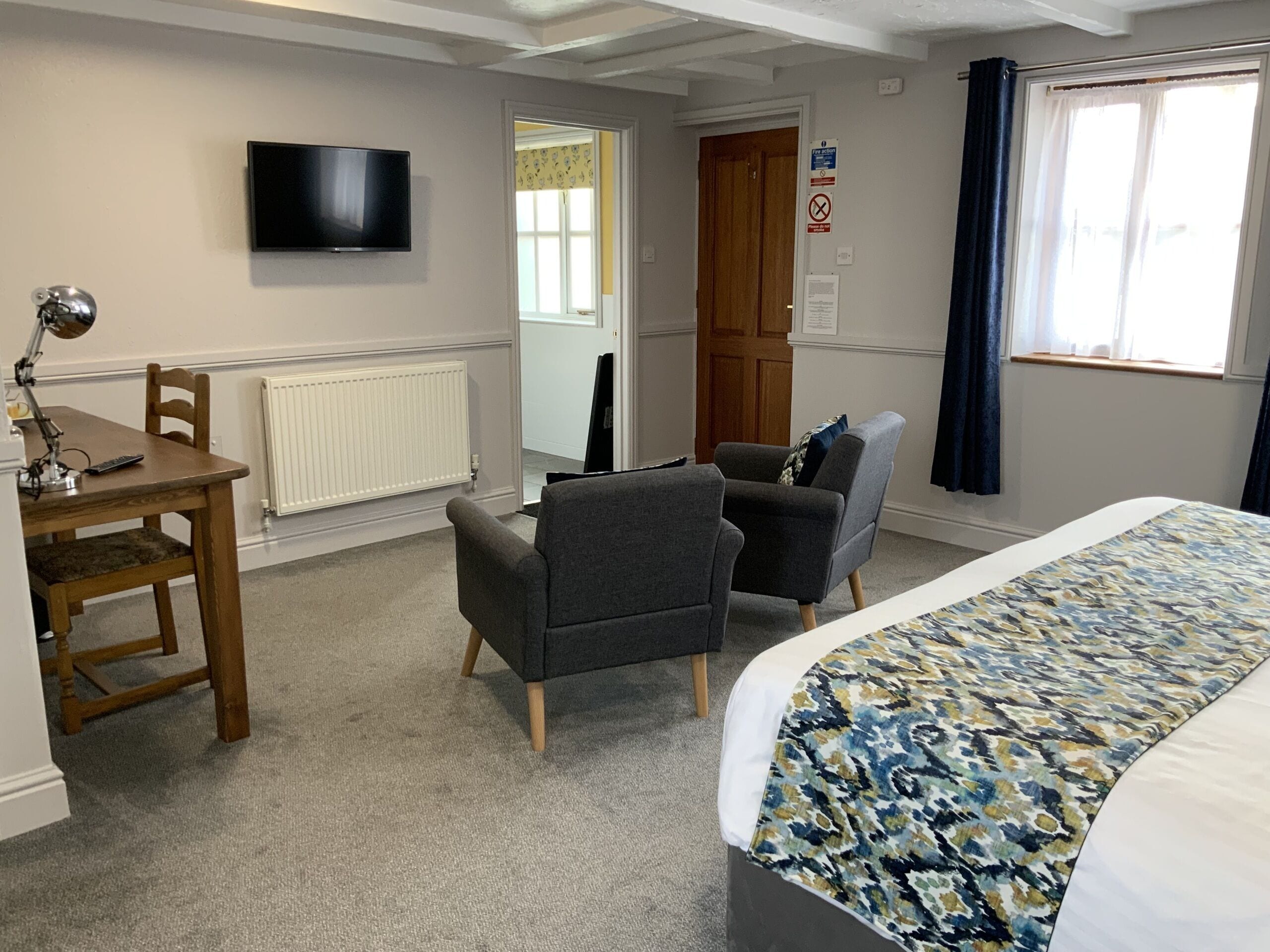 A hotel room with a bed, two armchairs, a desk with a lamp, a television, a radiator, and a window with curtains.
