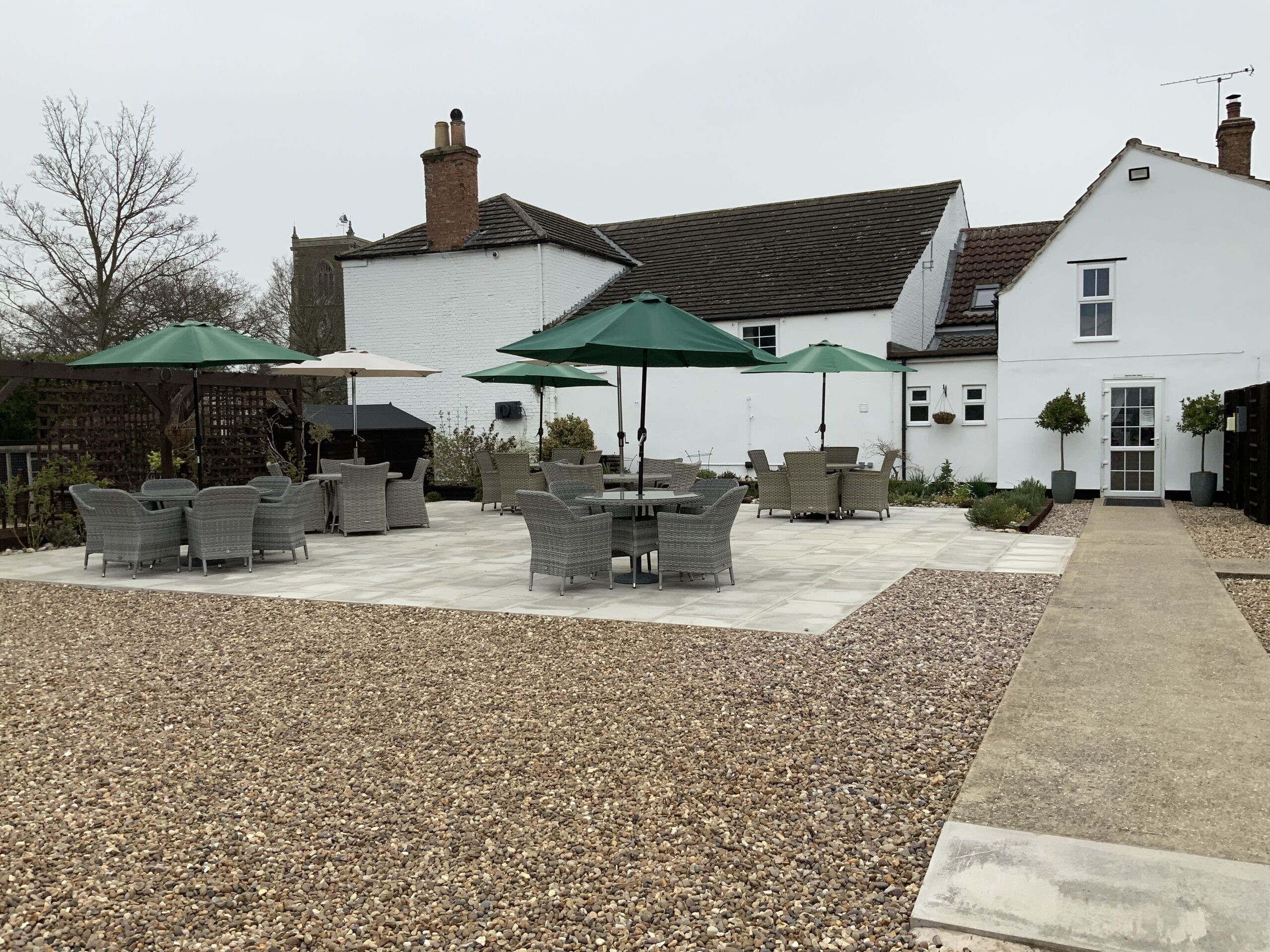 An outdoor patio area with wicker furniture and green umbrellas, a white building with a tiled roof, and a gravel path leading to the entrance.