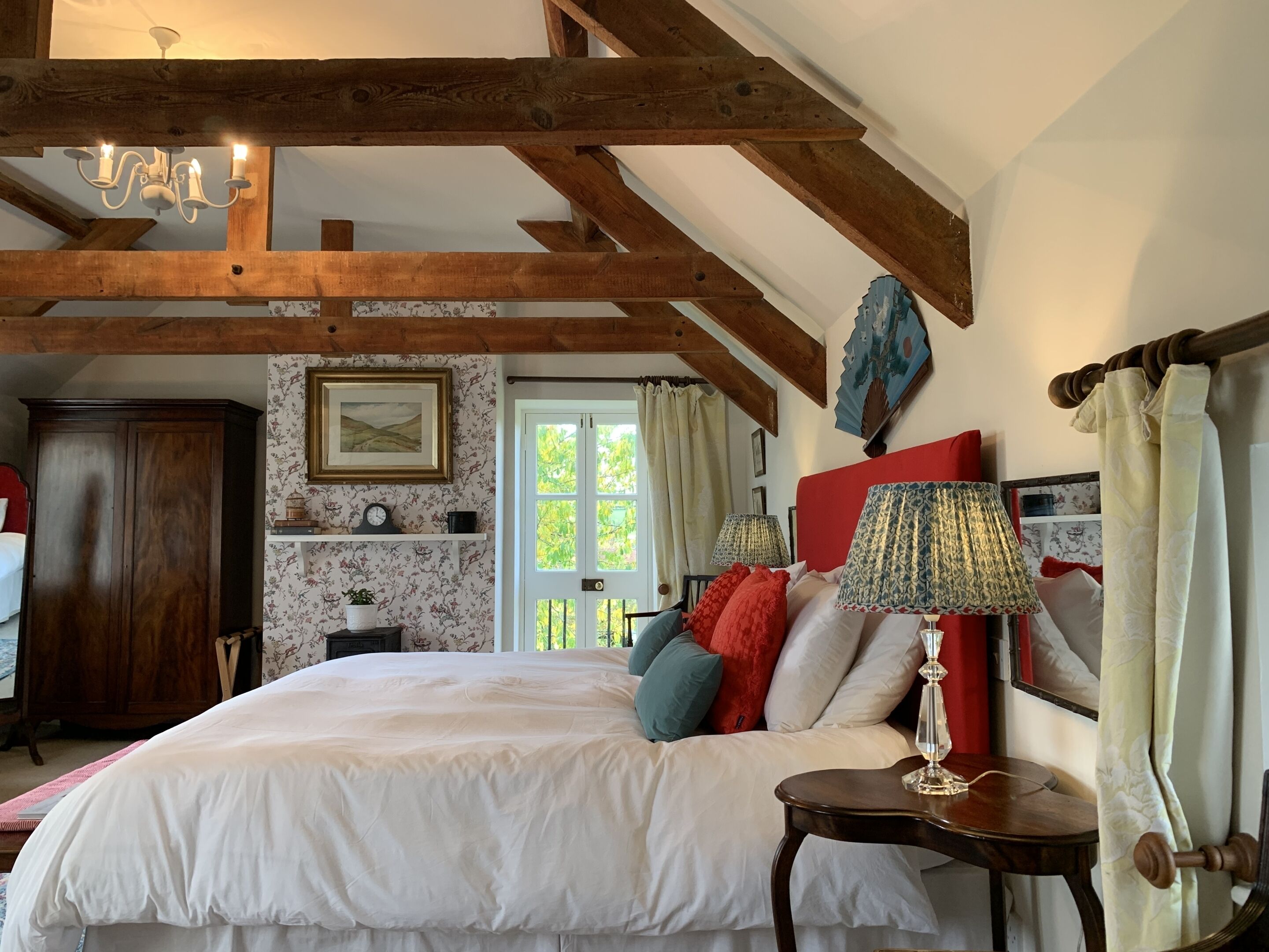Quarto com uma cama grande, vigas de madeira, um candelabro, uma mesa de cabeceira com abajur e uma janela com cortinas.