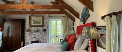 Quarto com uma cama grande, vigas de madeira, um candelabro, uma mesa de cabeceira com abajur e uma janela com cortinas.