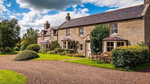 A stone house with a gravel driveway, a lawn, and a garden.