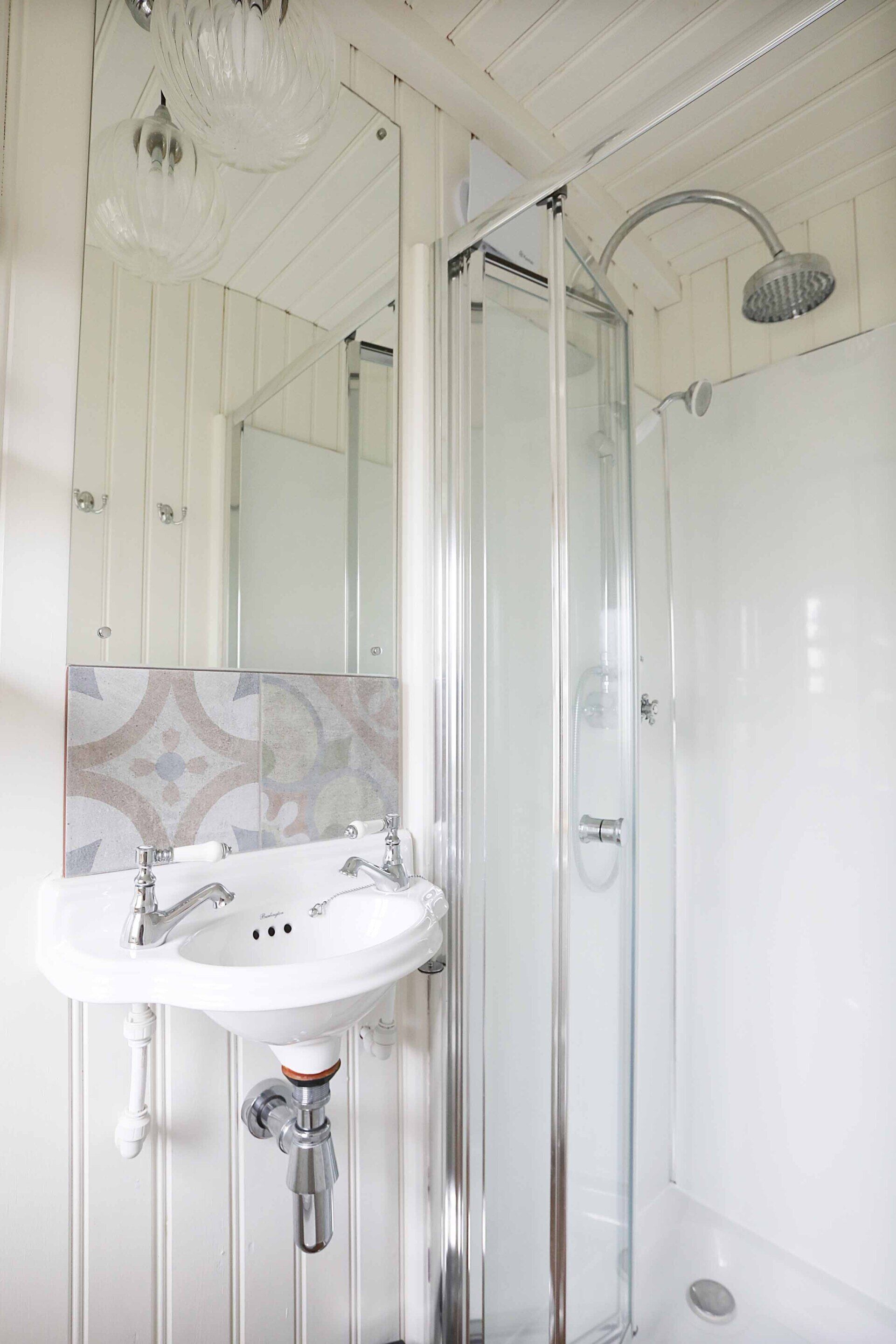 Une salle de bain dotée d’une douche à parois de verre, d’un lavabo blanc avec des robinets chromés et d’un mur recouvert de carreaux décoratifs.
