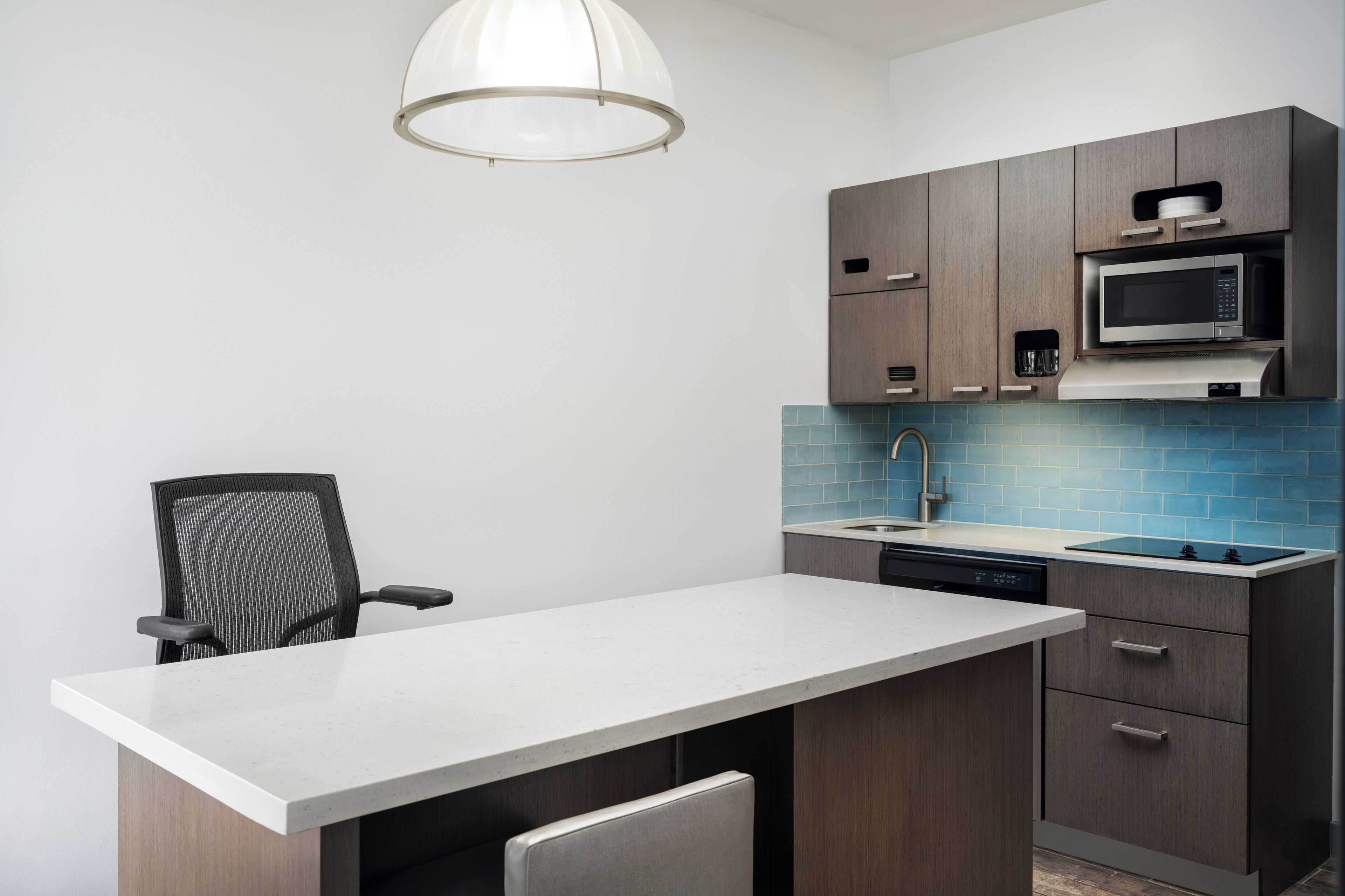 A modern kitchen with a white countertop, dark wooden cabinets, a built-in microwave, and a blue tiled backsplash.