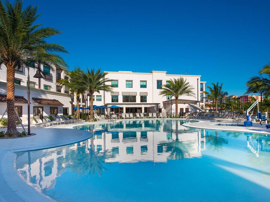 A pool area with lounge chairs and palm trees, surrounded by a building with multiple floors and balconies.
