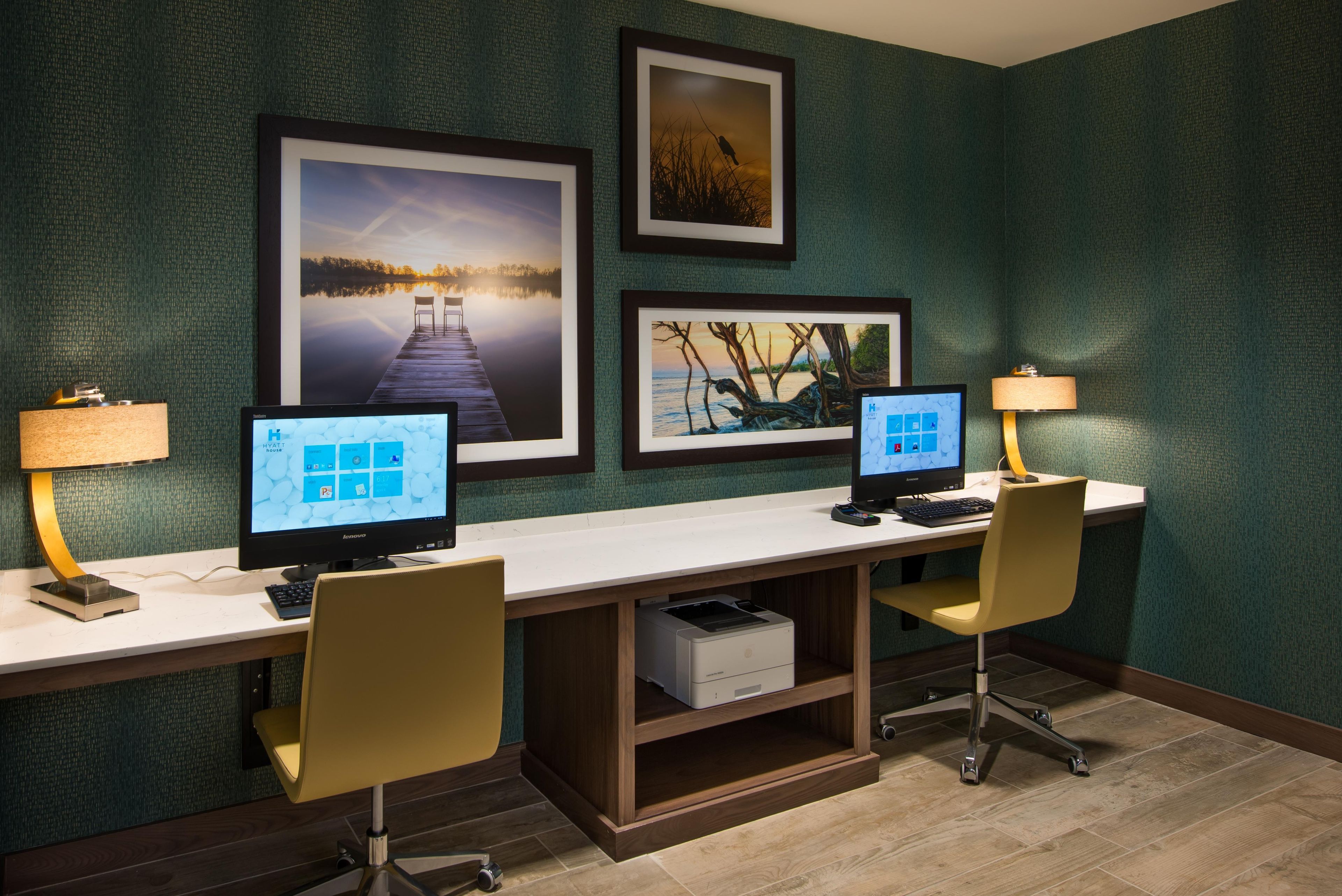 A hotel room with two desks, each with a computer and a lamp, framed pictures on the wall, and a wooden cabinet below the desks.