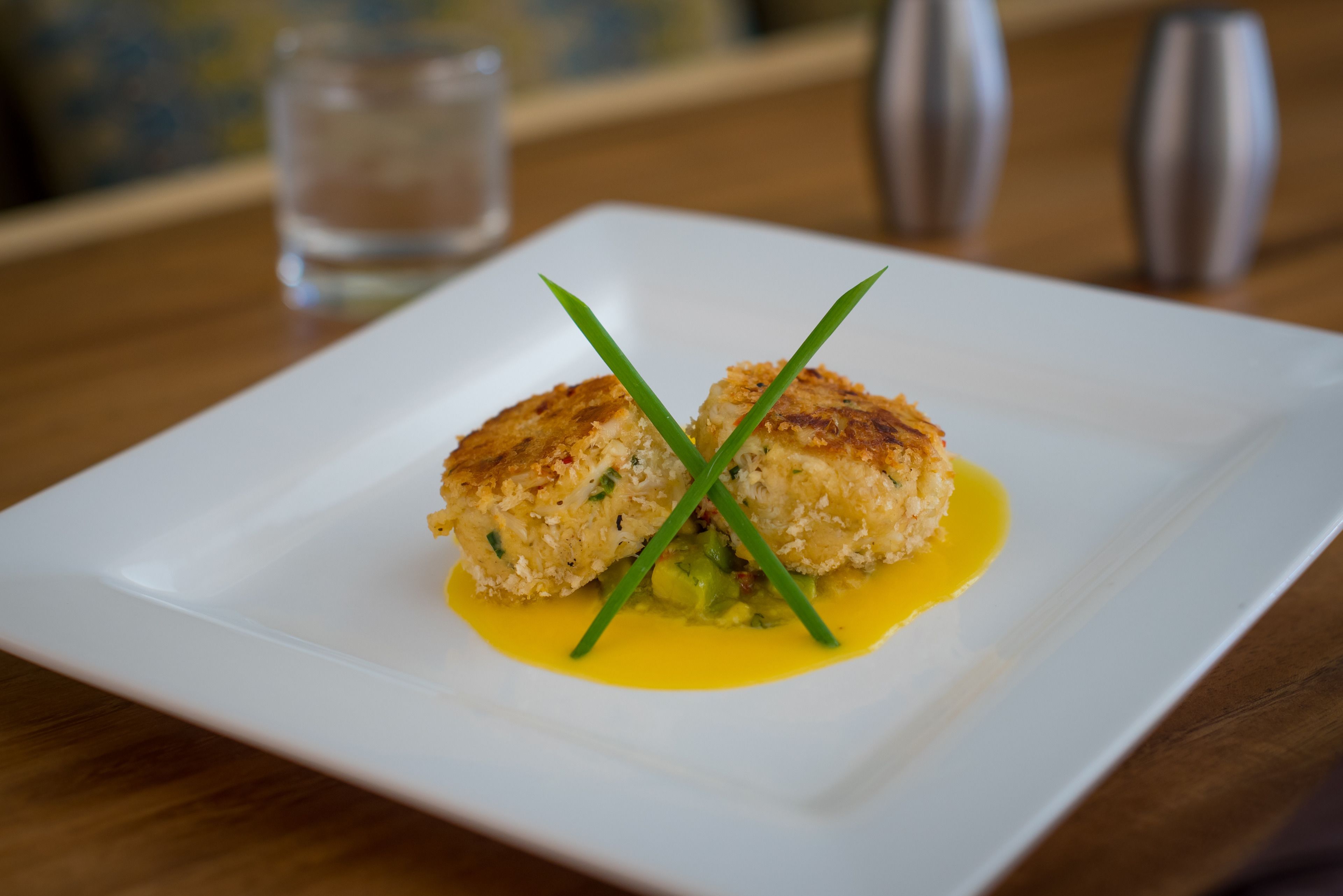 A square white plate with two golden-brown crab cakes garnished with green chives, served on a wooden table.