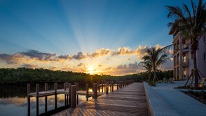 A wooden deck extending over water with a sunset in the background, a palm tree, and a building with balconies.