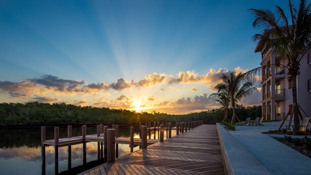 A wooden deck extending over water with a sunset in the background, a palm tree, and a building with balconies. - Hyatt House Naples/5th Avenue (Naples)