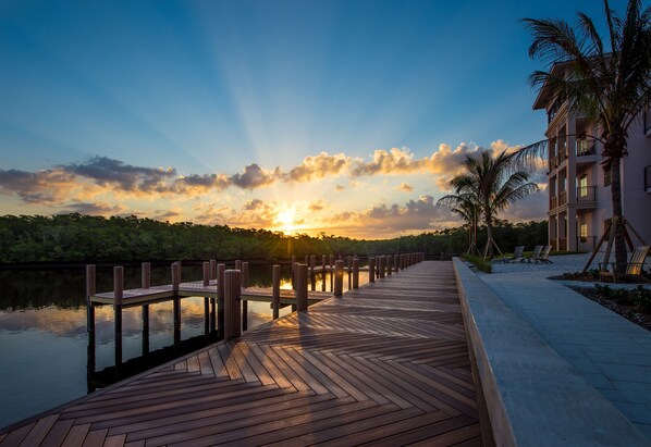 Um deck de madeira que se estende sobre a água, com um pôr do sol ao fundo, uma palmeira e um edifício com varandas.