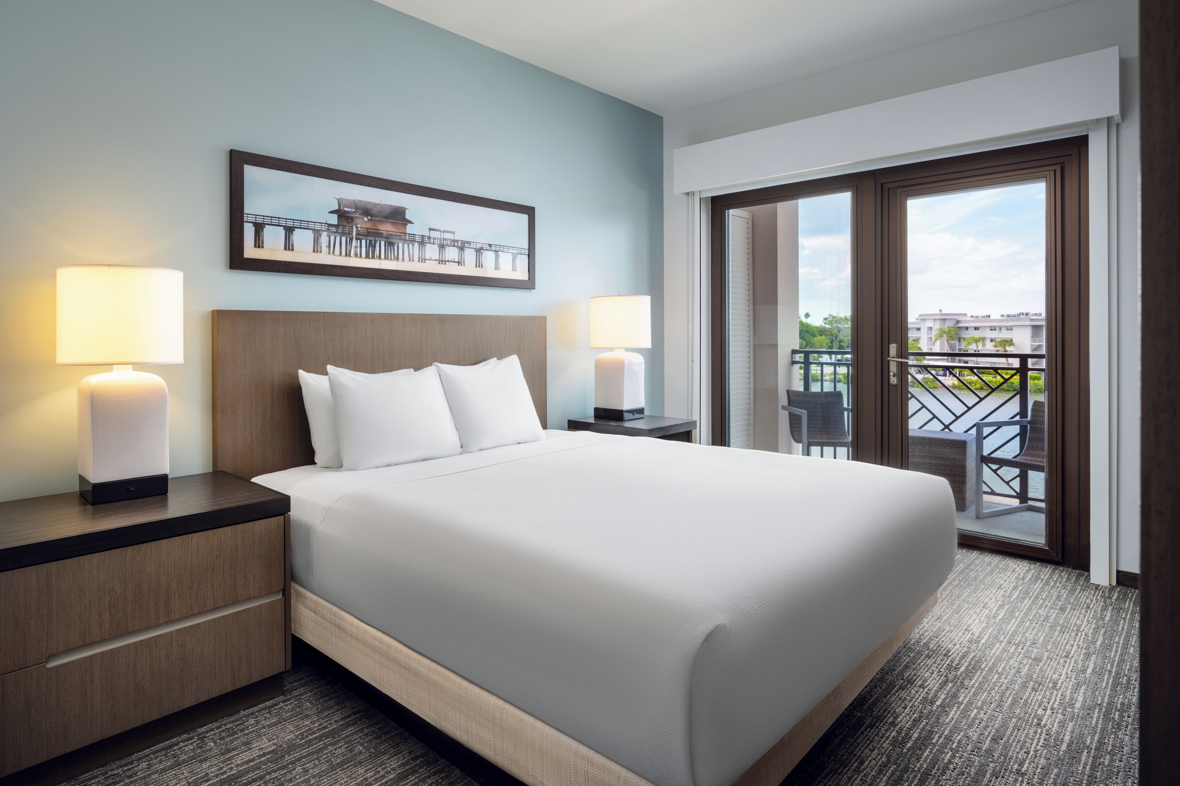 A hotel room with a large bed, two bedside tables with lamps, a view of the outdoors through a sliding door, and a framed picture on the wall.