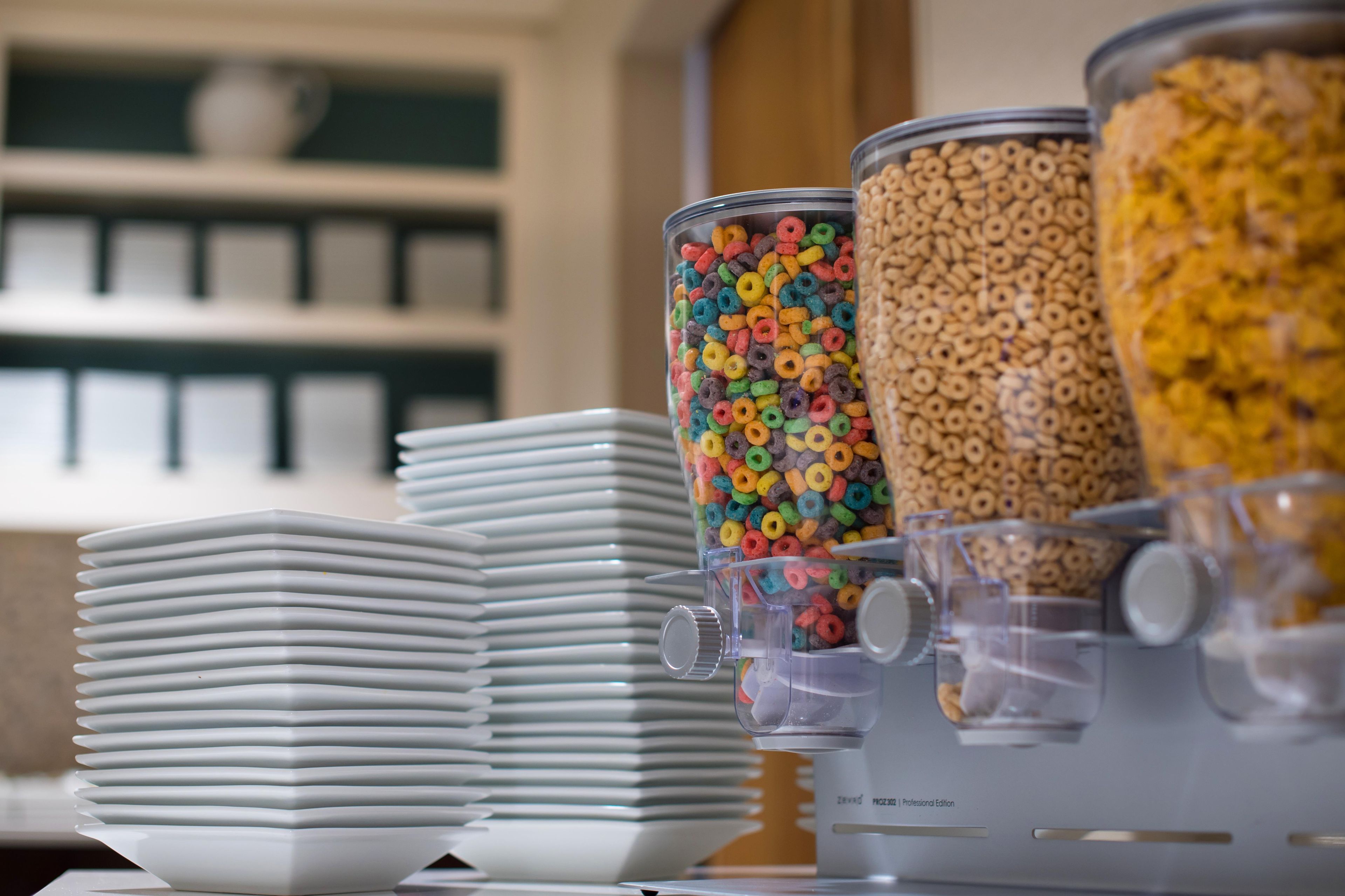 A cereal dispenser with multiple containers of colorful cereal and a stack of white plates.
