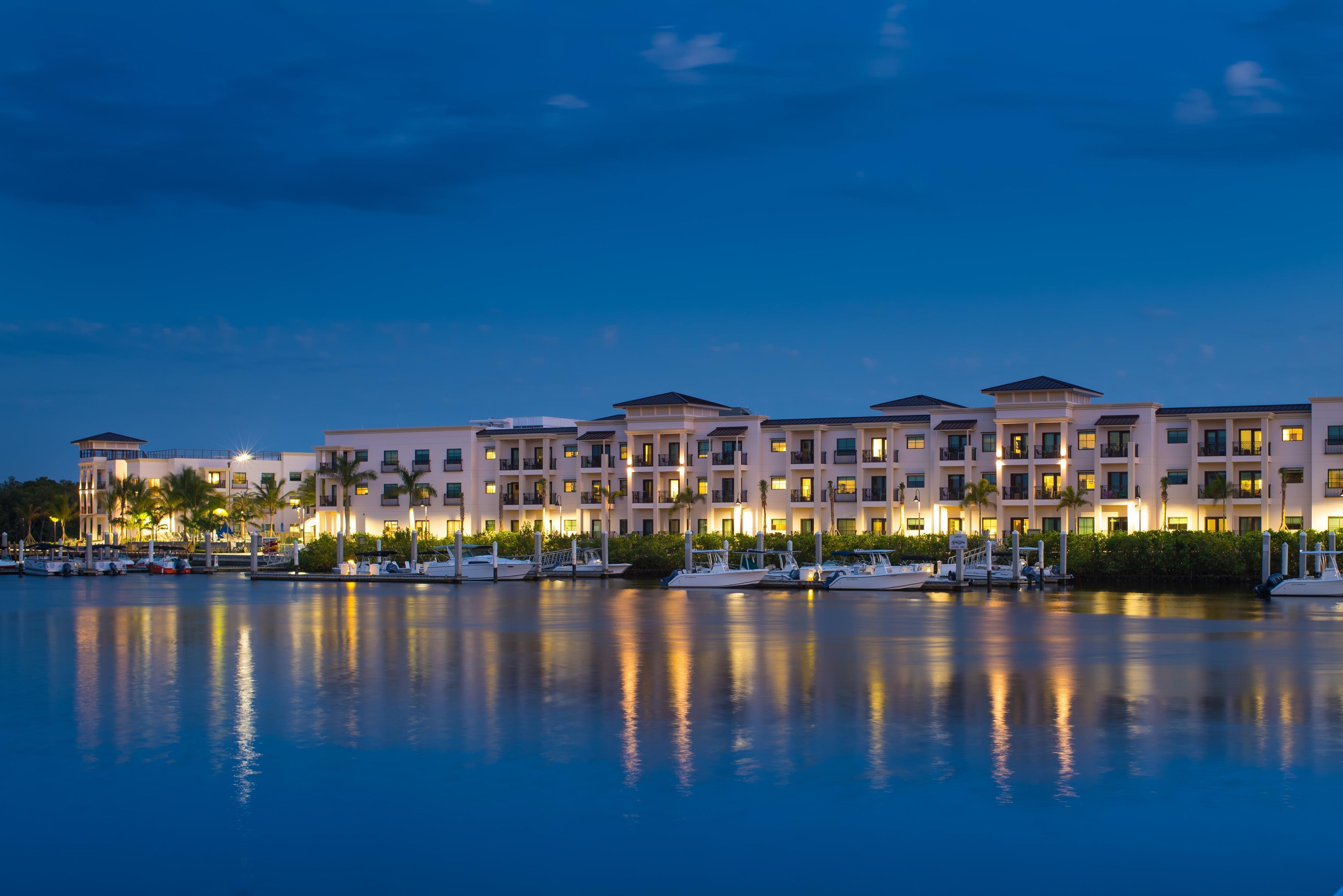 A waterfront hotel with multiple balconies, a marina, and a calm body of water reflecting the buildings.