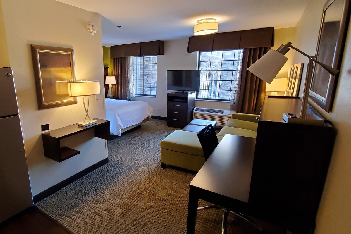 Habitación de hotel con cama, escritorio, refrigerador, televisor y vista a la ciudad.