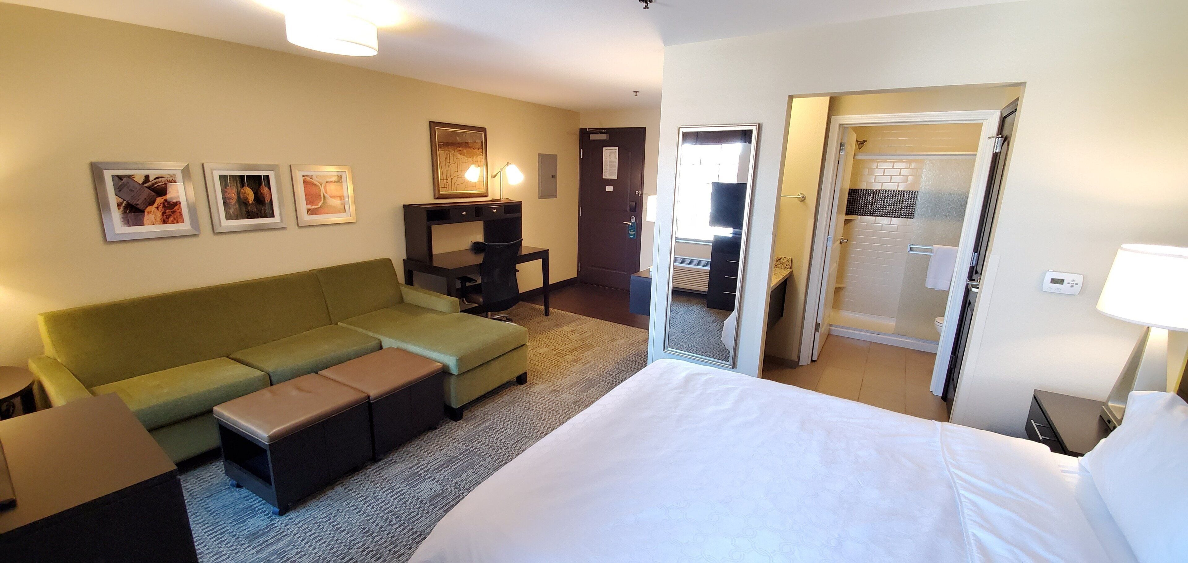 A hotel room with a bed, a green sofa, a desk with a chair, and a bathroom visible through an open door.