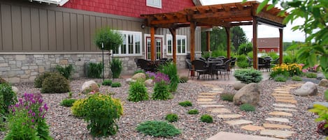 A red barn-style building with a stone foundation, a wooden pergola with outdoor seating, and a landscaped garden with various plants and a stone pathway.