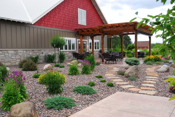 A red barn-style building with a stone foundation, a wooden pergola with outdoor seating, and a landscaped garden with various plants and a stone pathway.