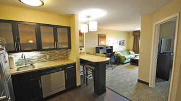 A modern kitchen with dark cabinets, a granite countertop, and stainless steel appliances. There is a dining area with a table and chairs, and a living room with a couch and a flat-screen TV.
