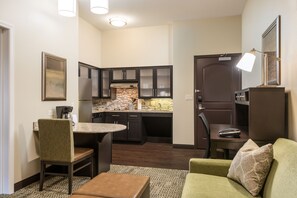 Un espacio habitable compacto con kitchenette, una zona de comedor con una mesa redonda y dos sillas, y un sofá verde con un cojín estampado.