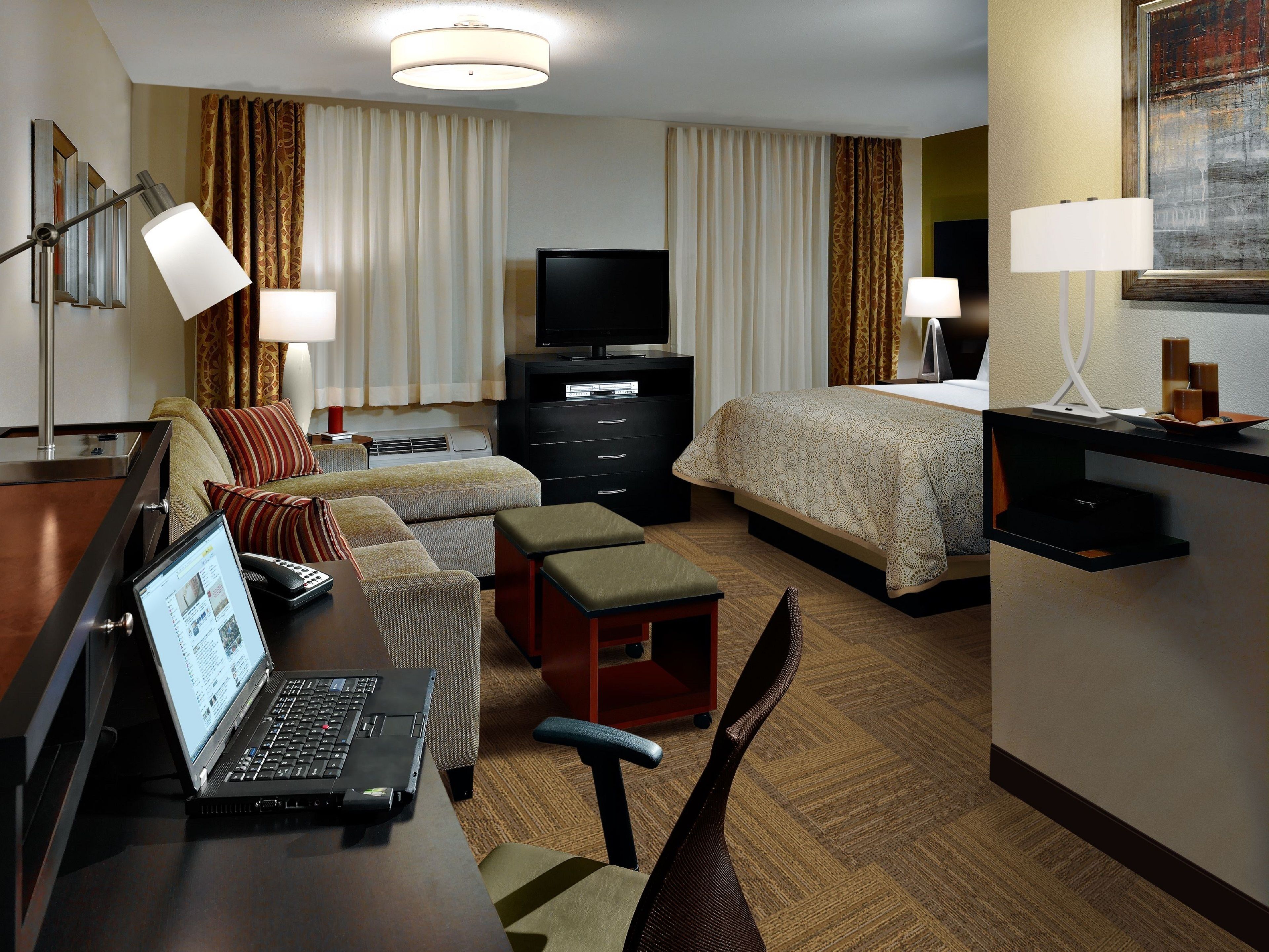 Habitación de hotel con cama, escritorio, laptop, televisor, cómoda y silla.