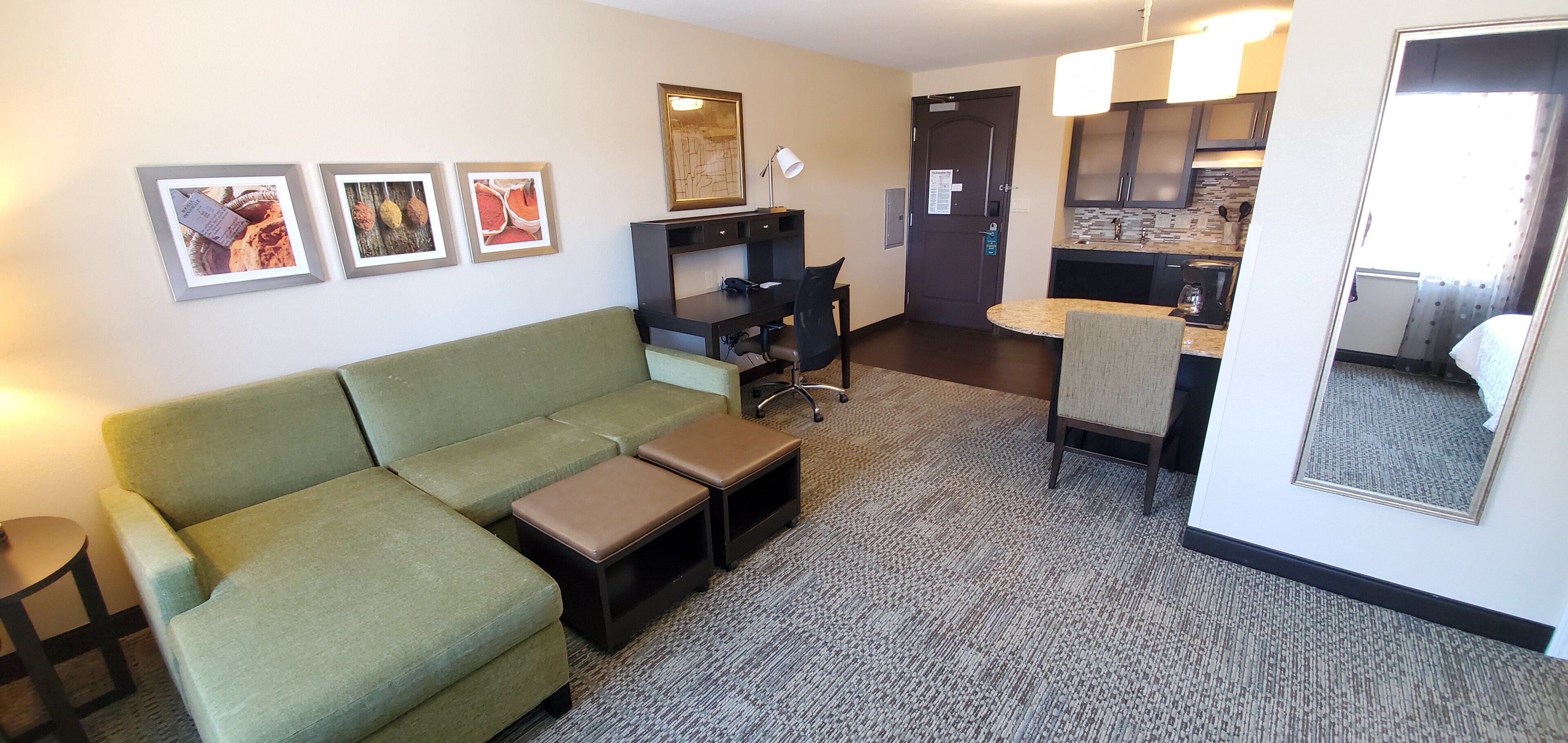 A hotel room with a green sofa, a small table, a desk with a chair, a kitchen area, and a bathroom.