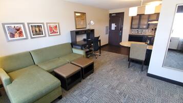 A hotel room with a green sofa, a small table, a desk with a chair, a kitchen area, and a bathroom.