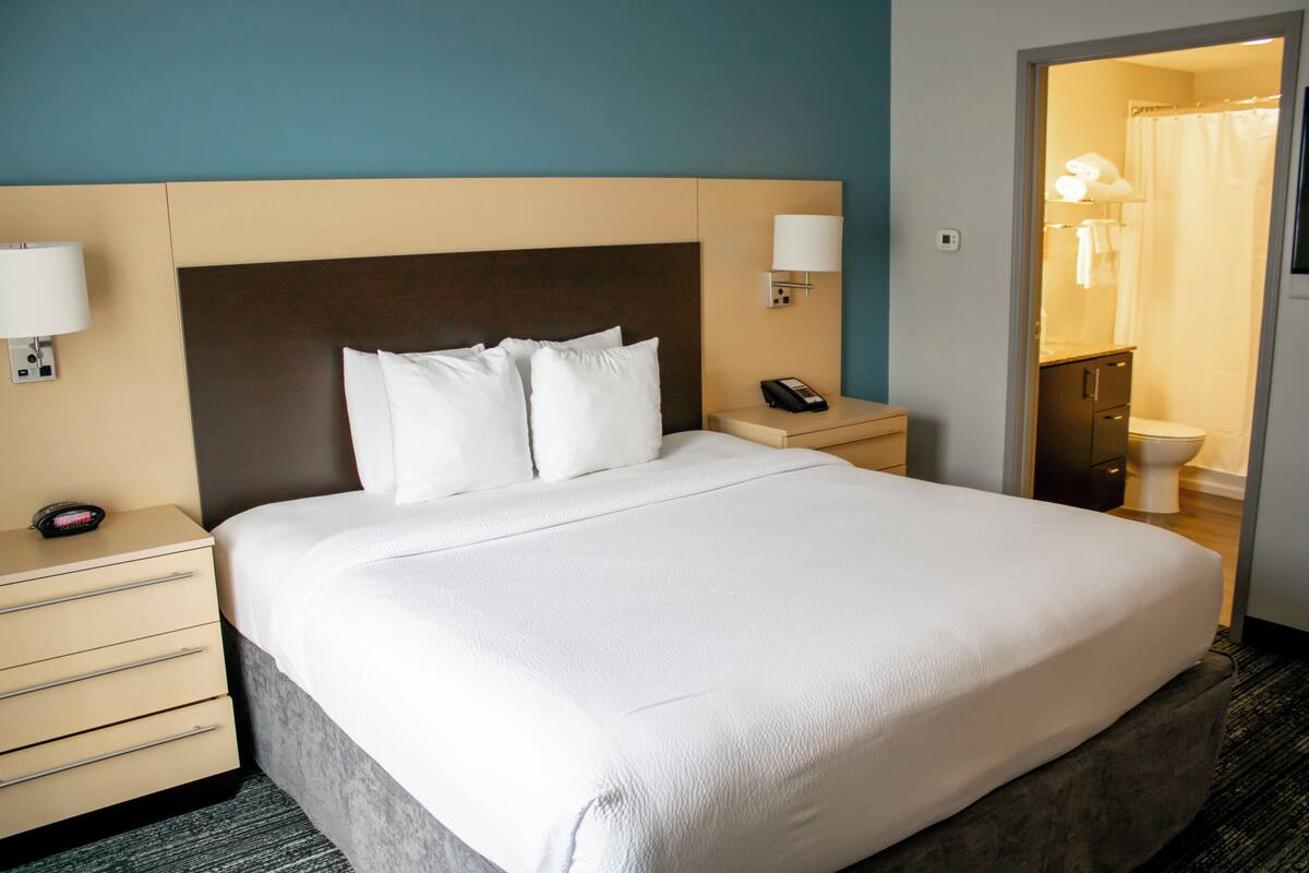 Ein Hotelzimmer mit einem großen Bett, Nachttischen, einer Lampe und Blick ins Badezimmer.