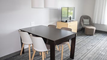 Une chambre d’hôtel avec une table à manger en bois foncé, des chaises blanches, un buffet beige, un téléviseur et un fauteuil gris.