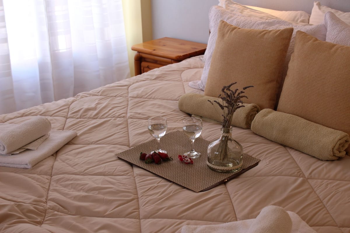 A neatly made bed with a beige quilt, two rolled towels, a tray with two glasses of water, a vase with dried flowers, and a wooden nightstand in the background.