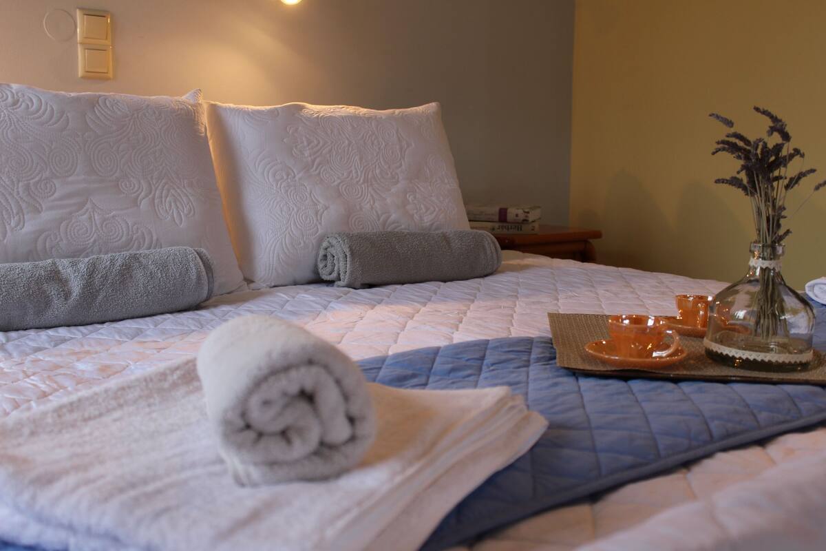 A neatly made bed with rolled towels, a blue quilt, and a tray with orange cups and a vase of dried flowers.