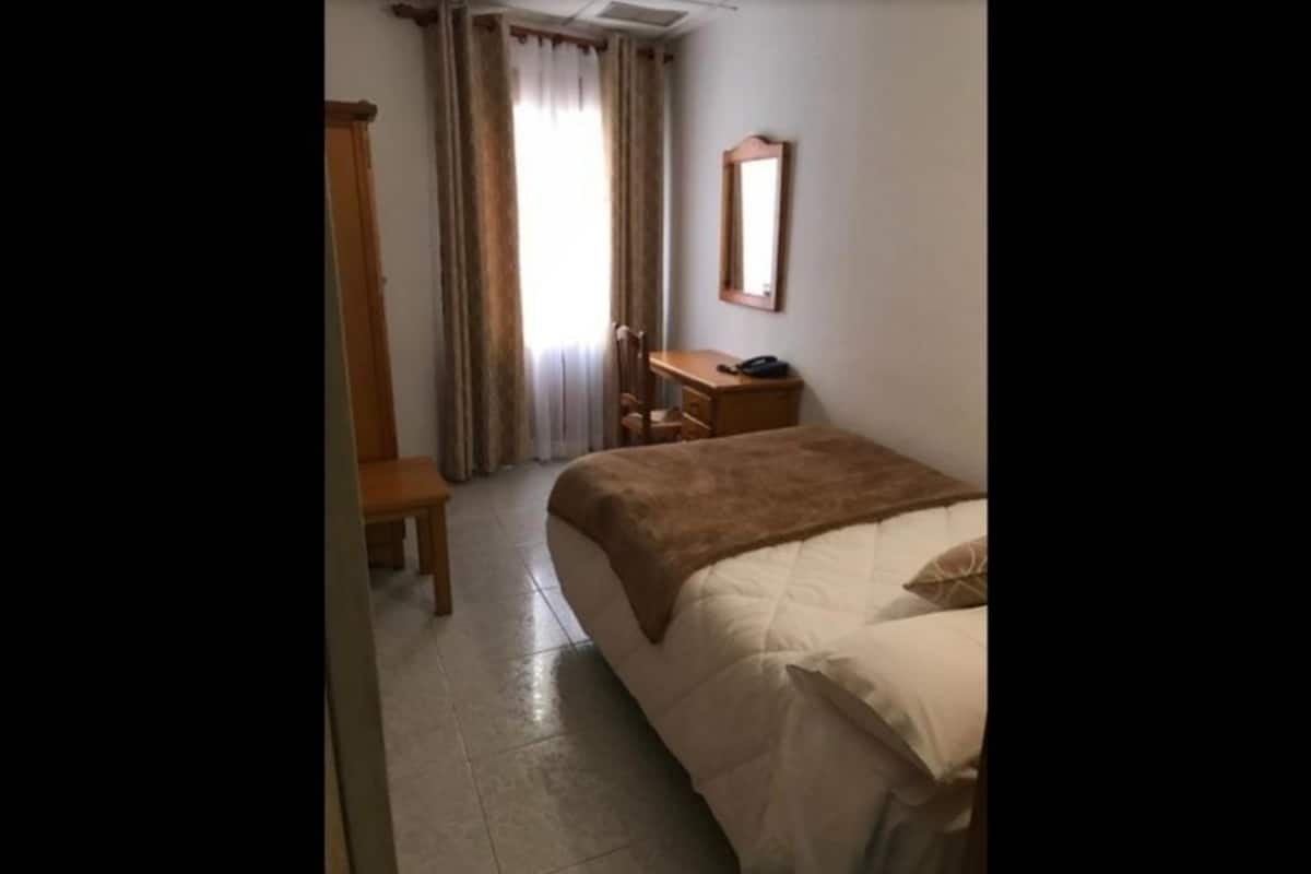 A hotel room with a bed, a desk, a chair, a mirror, and a window with curtains.