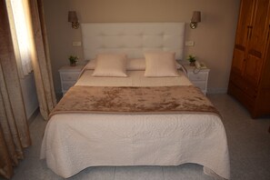 A neatly made bed with a white headboard and two pillows, a bedside table with a lamp, and a wooden wardrobe. - Hostal Alcázar (Toledo)