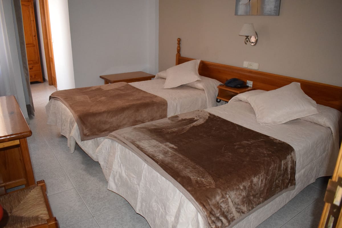 A hotel room with two beds, a wooden headboard, a bedside table with a lamp, and a small desk with a chair.