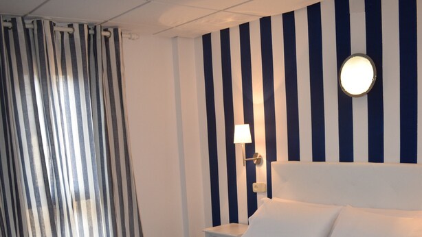 A neatly made bed with a dark blue blanket, a striped curtain, and a wall with vertical stripes and a round light fixture. - Hostal Alcázar (Toledo)