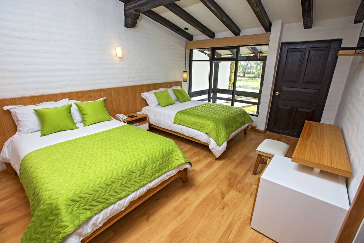 Habitación con dos camas, cada una con ropa de cama verde, cabeceros de madera y almohadas blancas. Hay una mesita de madera y un banco.