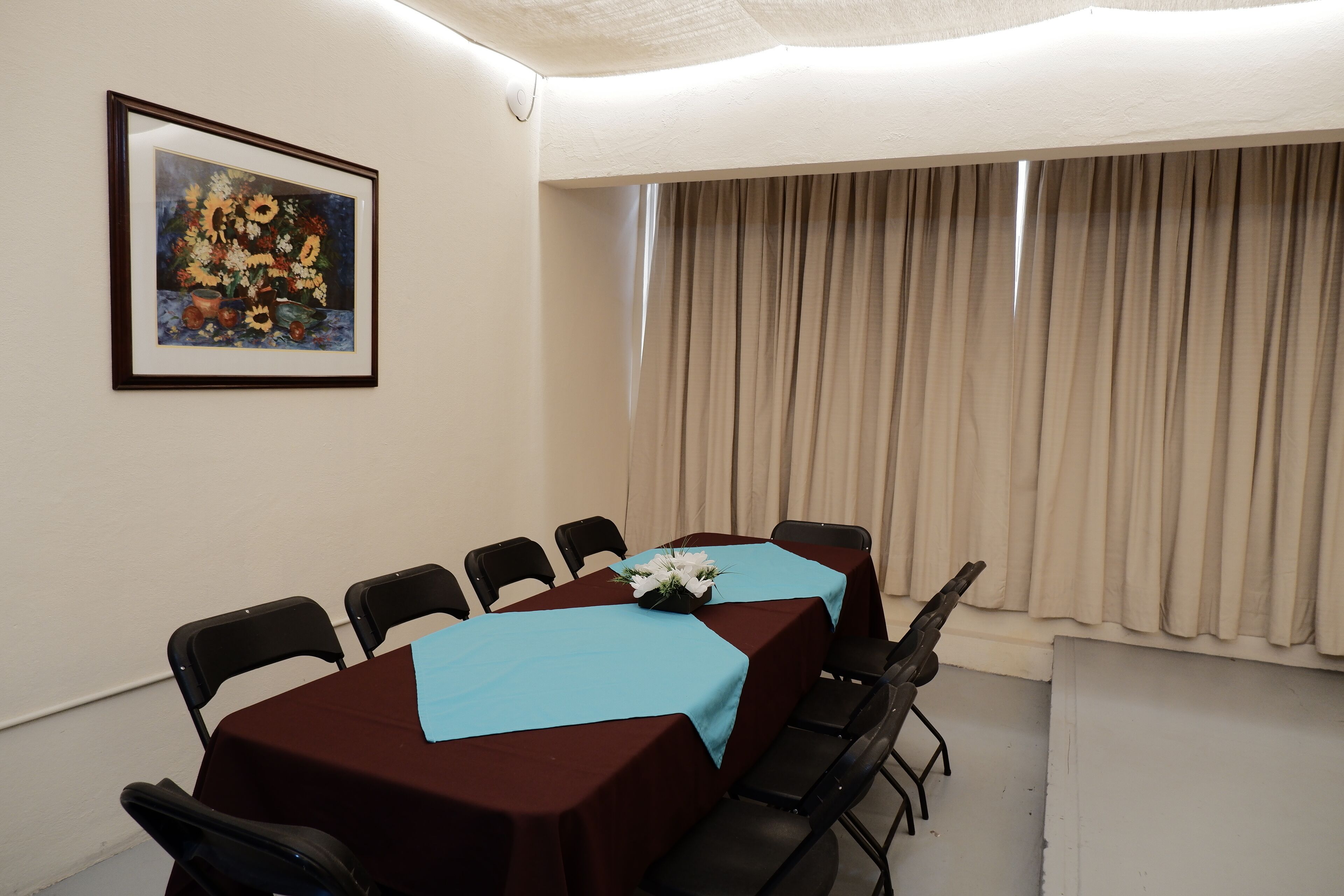 Un salón de conferencias con una larga mesa, sillas negras, mantel azul y un cuadro enmarcado en la pared.