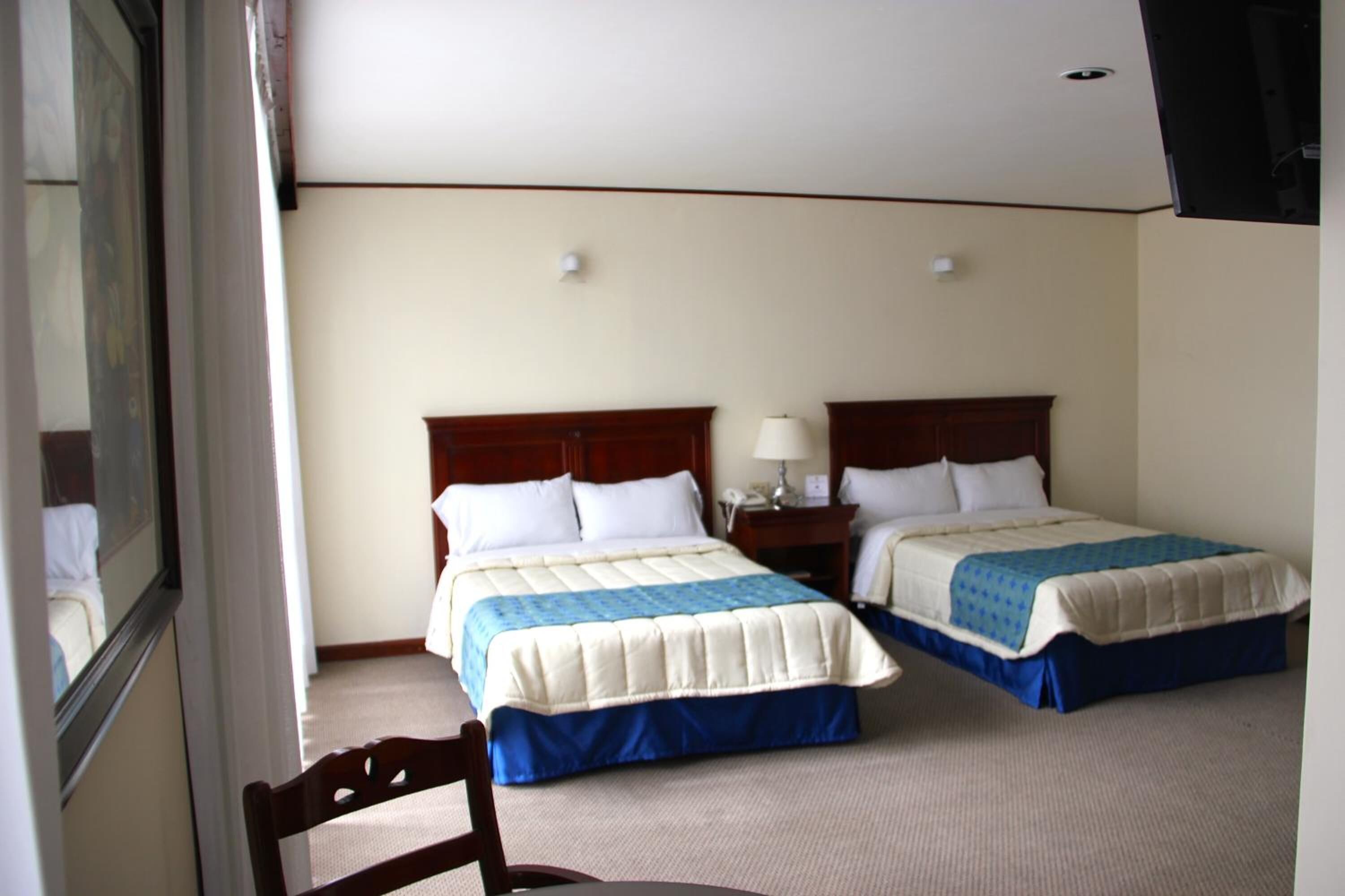 A hotel room with two beds, a chair, a nightstand with a lamp, and a window.