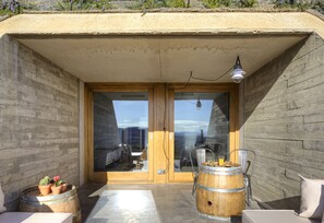 A modern outdoor seating area with wooden barrels, a glass door entrance, and a view of the cityscape. - Trossos del Priorat (Gratallops)