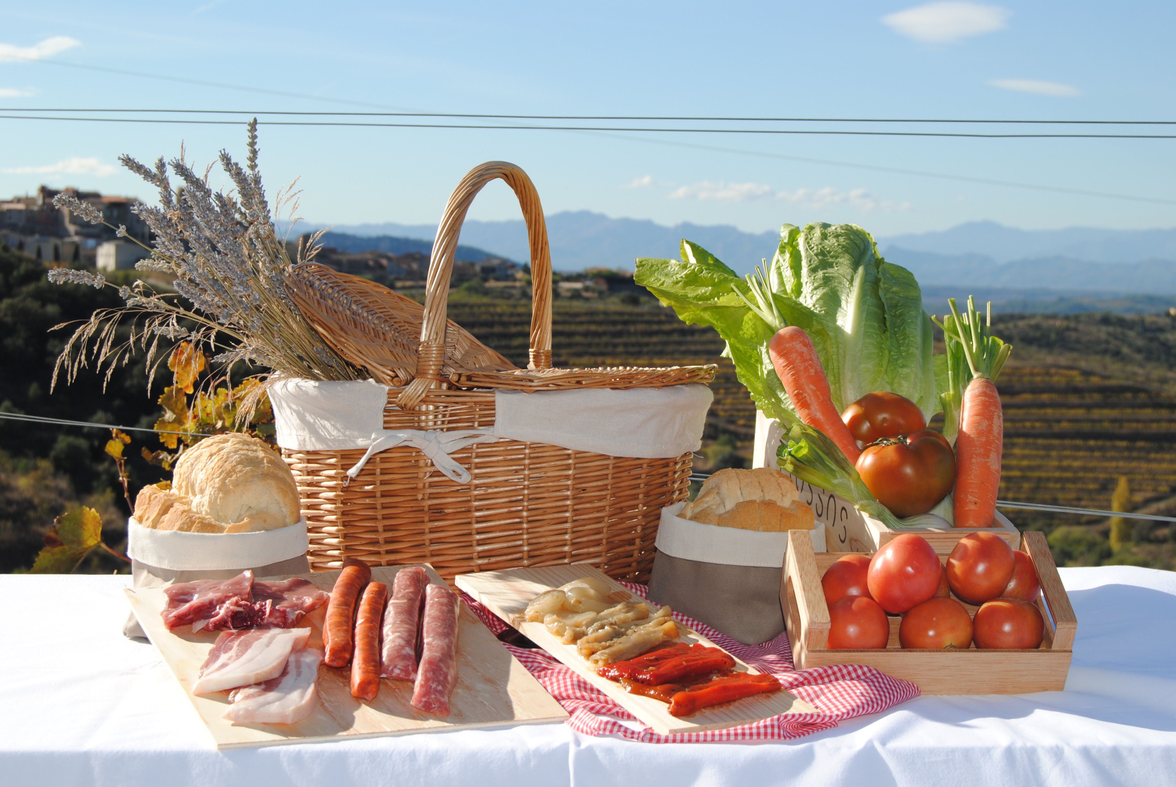 Un àpat de pícnic amb pa, carns, verdures i una cistella sobre una taula amb una vista panoràmica.
