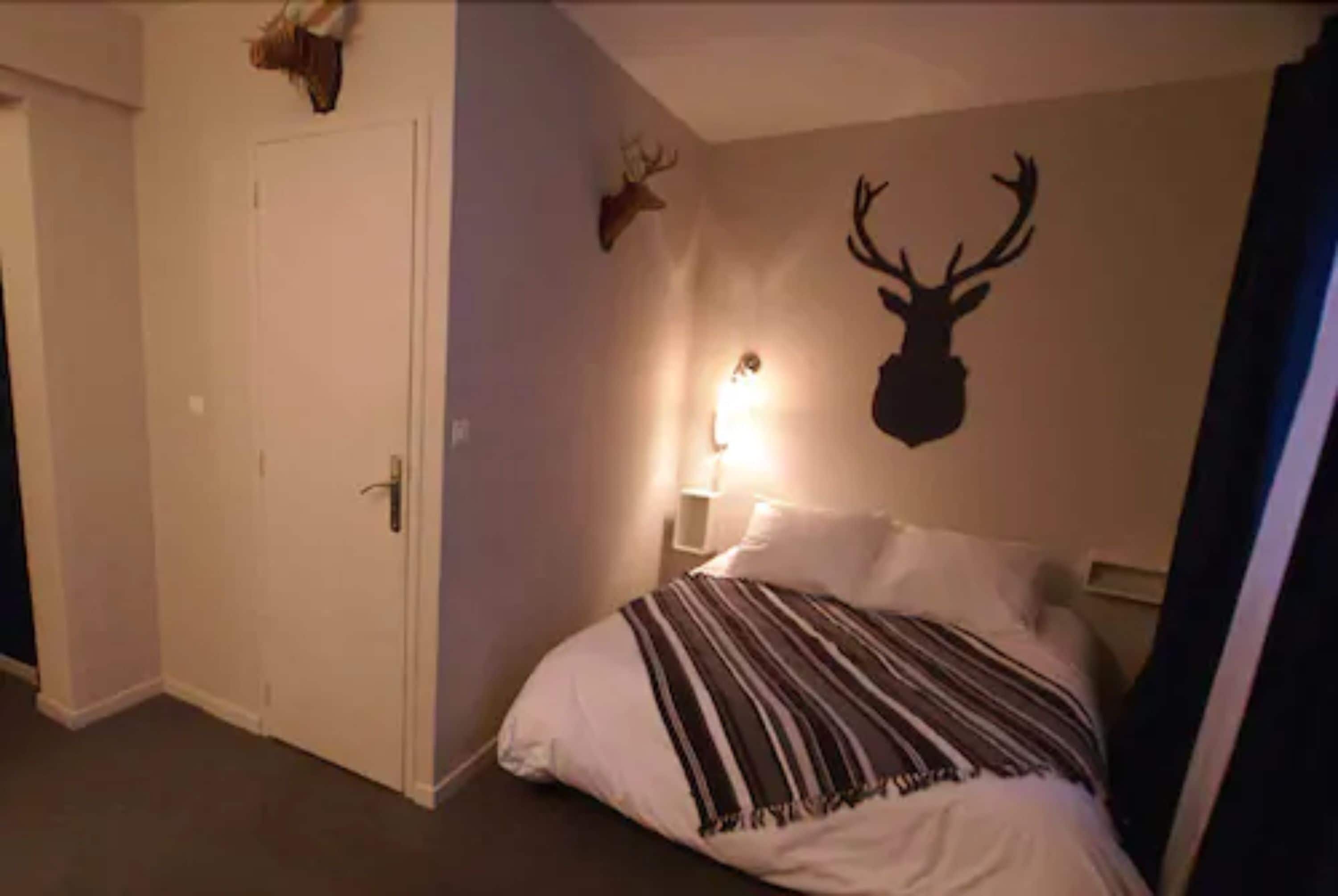 Une chambre à coucher avec un lit, une décoration murale représentant une tête de cerf, une porte et une table de chevet avec une lampe.