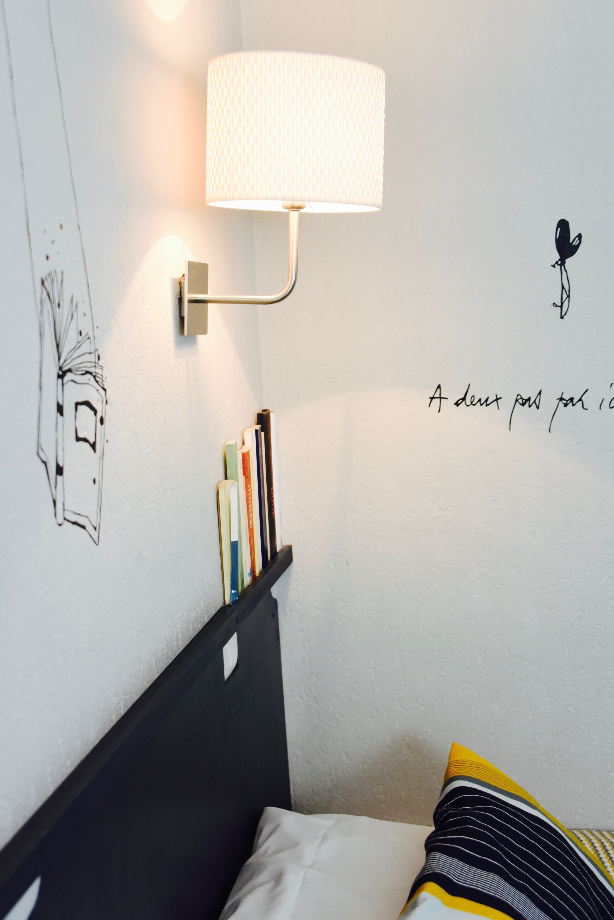 A neatly arranged bedroom with a bed, a bedside table with books, a wall-mounted lamp, and a wall decoration.
