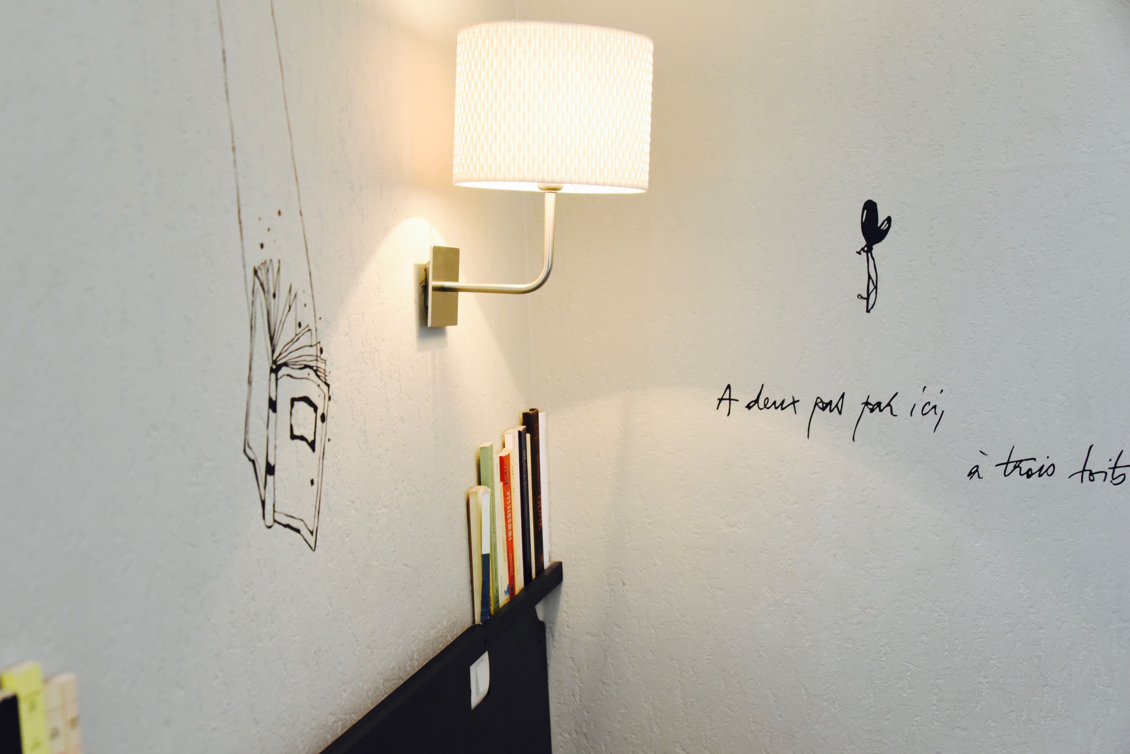 Una pared con una lámpara de pared, una estantería llena de libros y un adhesivo decorativo con texto e ilustración.