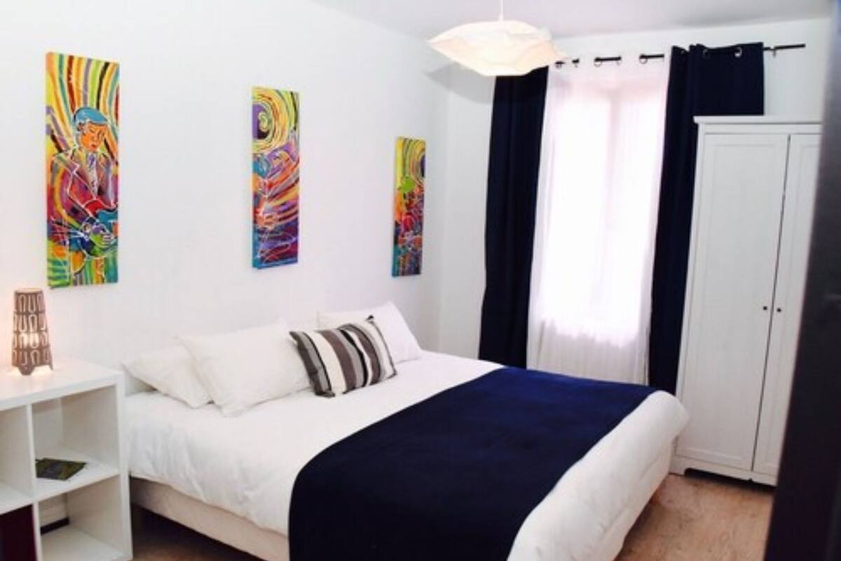 A bedroom with a bed, white walls, colorful abstract paintings, a wardrobe, and a bedside table with a lamp.