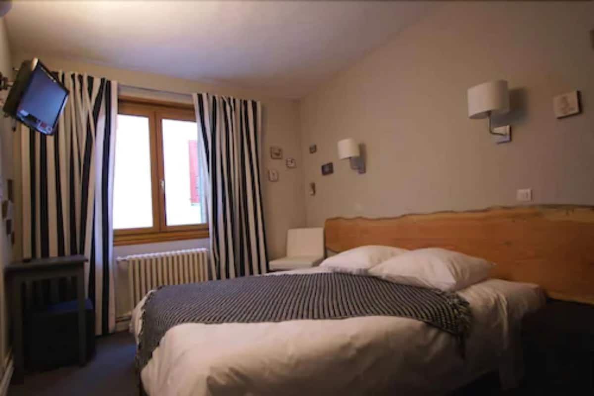 A hotel room with a bed, a TV mounted on the wall, a window with striped curtains, and a radiator.