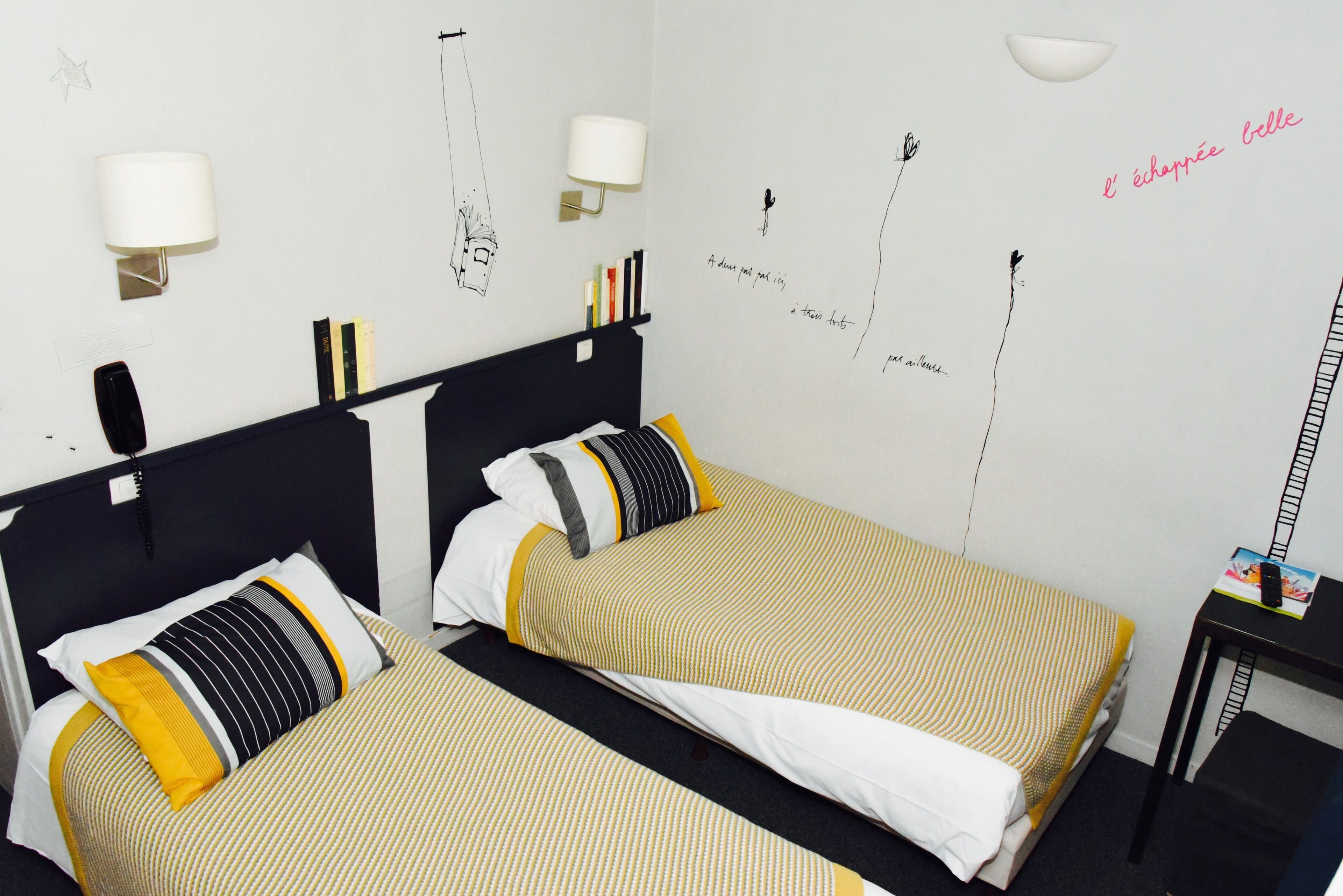 Camera d'albergo con due letti, ciascuno dotato di una testiera nera e biancheria a righe gialle. Sono presenti lampade a parete e un tavolino con un libro e un telefono.